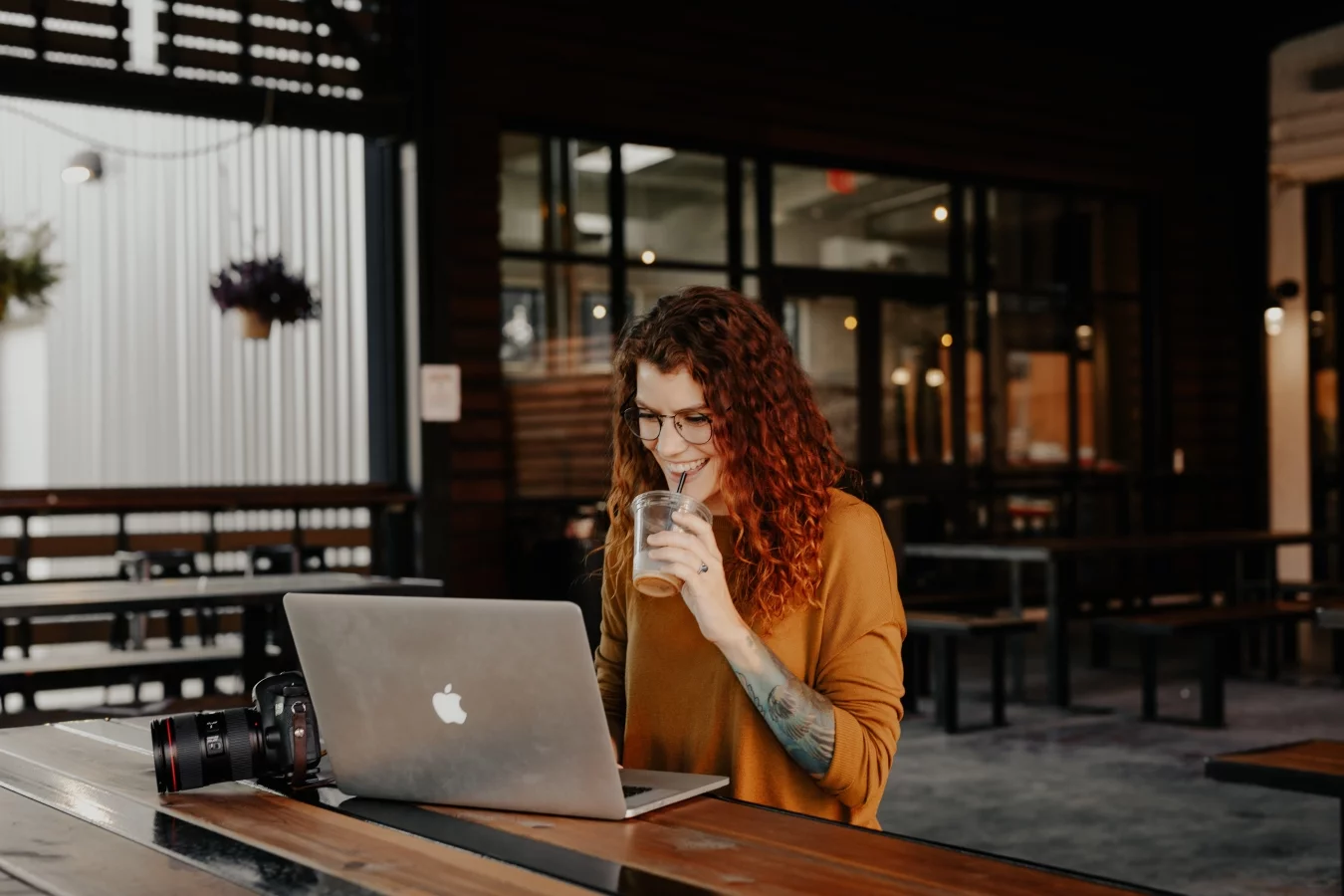 Red-haired woman sipping a drink while smiling at a MacBook, with a camera on the table, learning Shopify Liquid