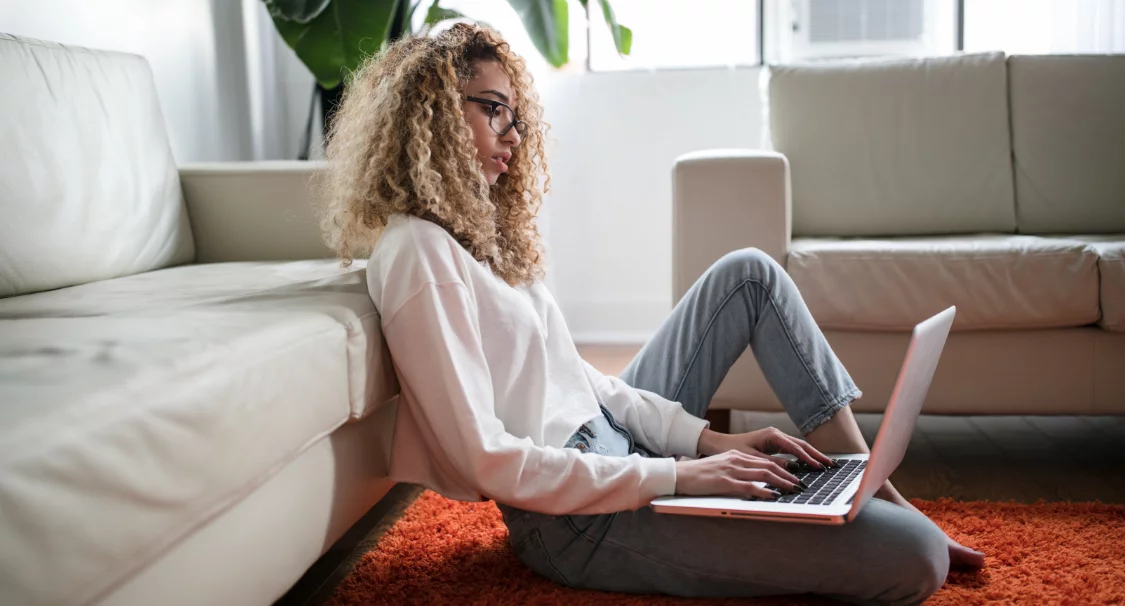 Woman with curly hair sitting on an orange rug against a sofa, working on a laptop before a Shopify migration