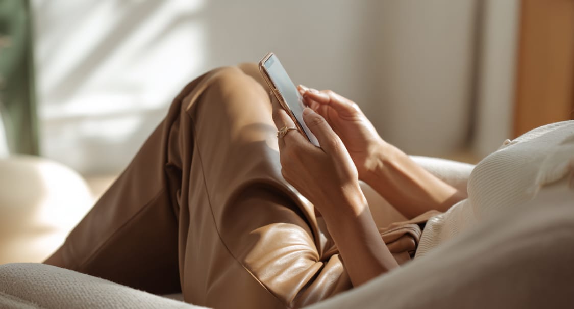 Kobieta w beżowych spodniach przegląda smartfon, siedząc wygodnie w fotelu w słonecznym wnętrzu