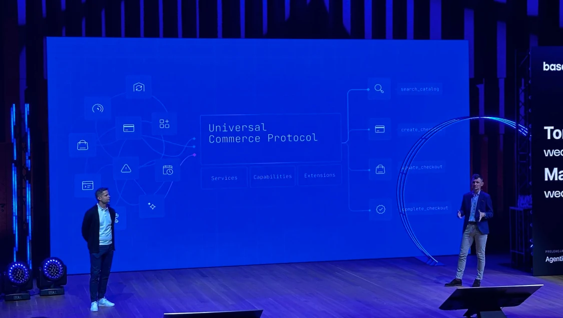 Two speakers on stage in front of a large blue screen presenting the 'Universal Commerce Protocol' diagram