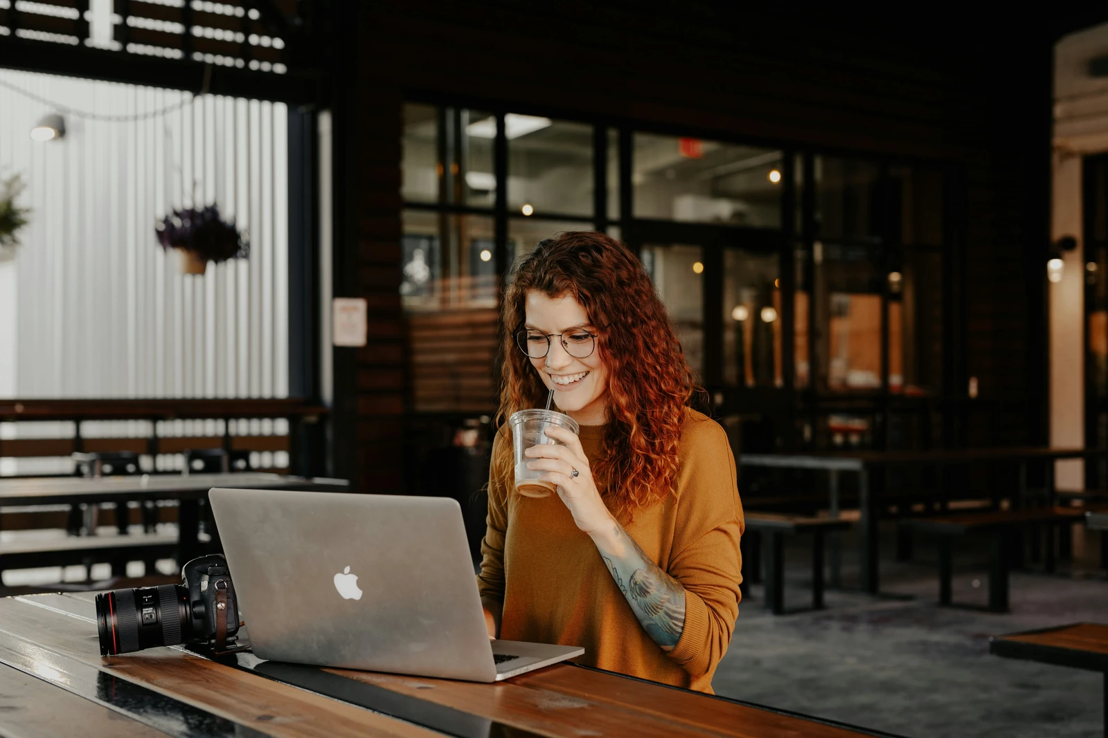 Smiling red-haired woman with a drink working on a laptop at a café table next to a camera, browsing product pages