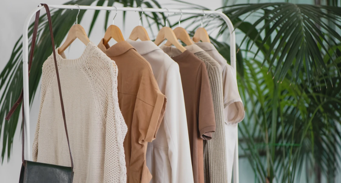 Wieszak z ubraniami w beżowych i brązowych odcieniach oraz skórzana torebka na tle palmy, nawiązujące do sklepu fashion