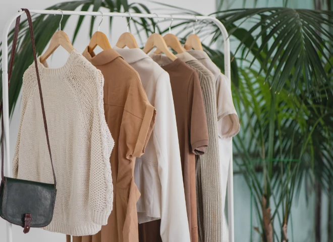 Wieszak z beżowymi i białymi ubraniami oraz skórzaną torebką na tle palmy, ilustrujący sklep fashion