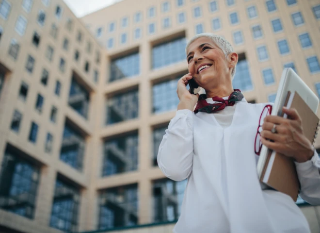 Uśmiechnięta siwowłosa kobieta rozmawia przez telefon przed biurowcem, trzymając laptopa i notes