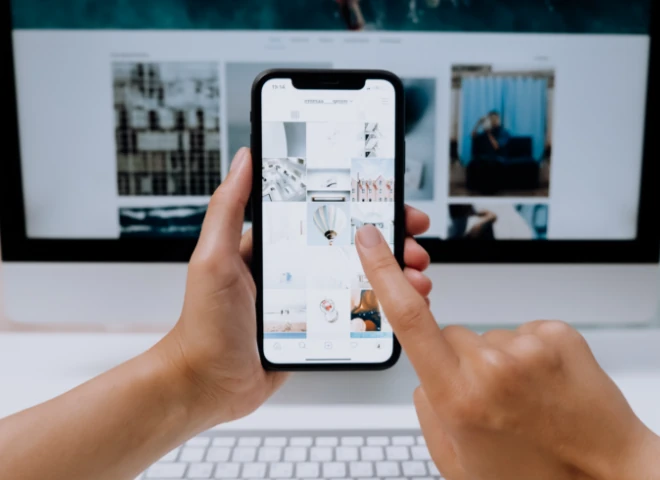 Dłonie przeglądają profil na Instagramie w smartfonie na tle monitora z galerią zdjęć