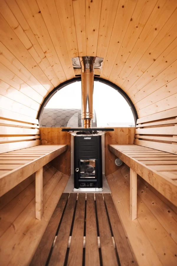 An inside image of a barrel sauna. We see a stove surrounded by a wooden decor.