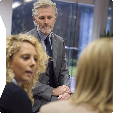 Older man in a suit attentively listening to two women during a business meeting in an office.