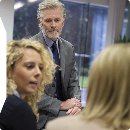 Older man in a suit attentively listening to two women during a business meeting in an office.