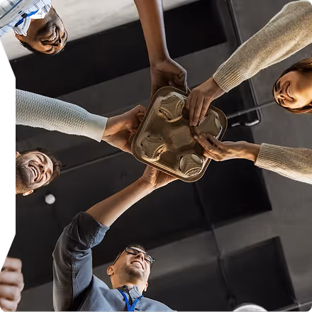 Four diverse people holding an empty cardboard drink carrier together, viewed from below.