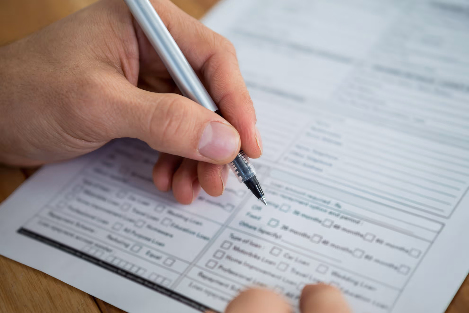 Hand holding a pen poised to fill out a detailed form with checkboxes and text fields on a wooden surface.
