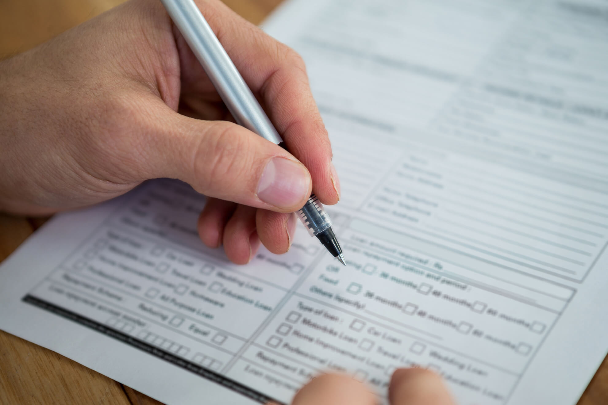 Hand holding a pen poised to fill out a detailed form with checkboxes and text fields on a wooden surface.