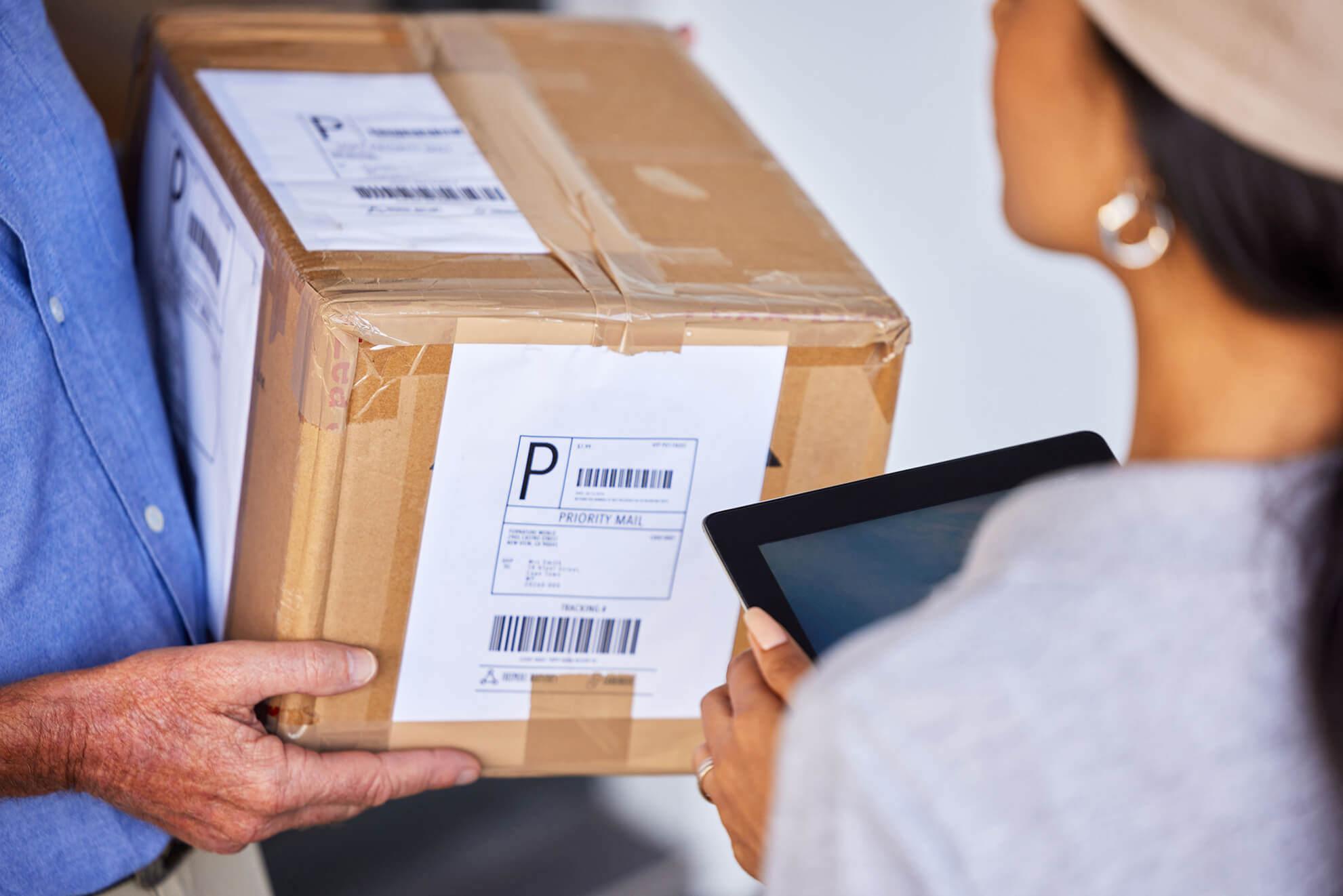 Person holding a cardboard package with a priority mail label, another person scanning it using a tablet device.