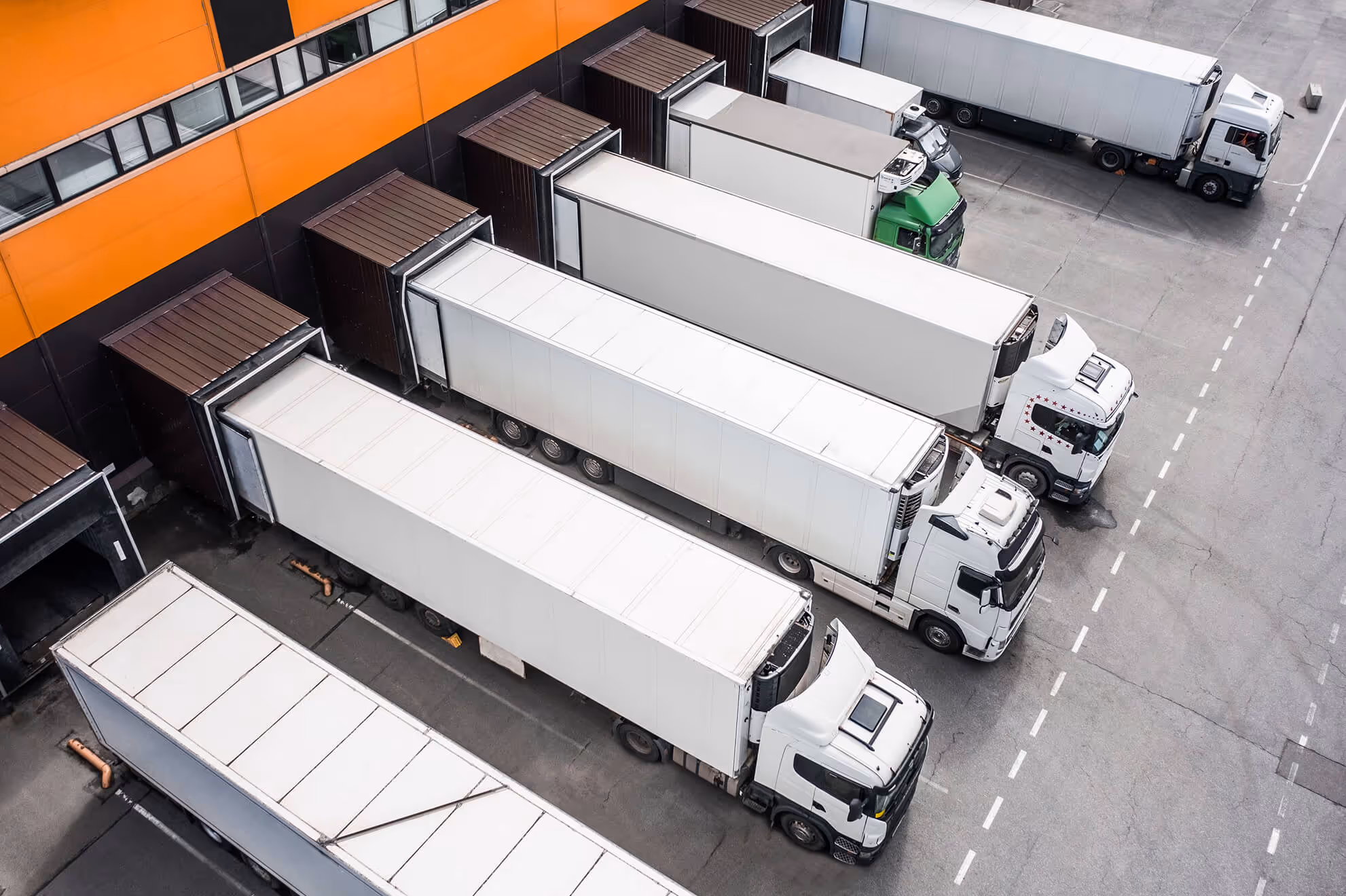 Aerial view of six white trucks docked at a loading bay attached to a building with orange and black walls.