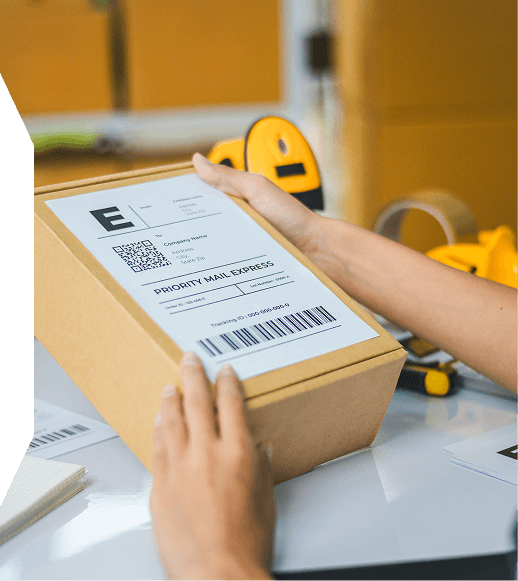 Hands holding a cardboard box with a Priority Mail Express shipping label on a white table, with packing tape dispenser and other boxes in the background.