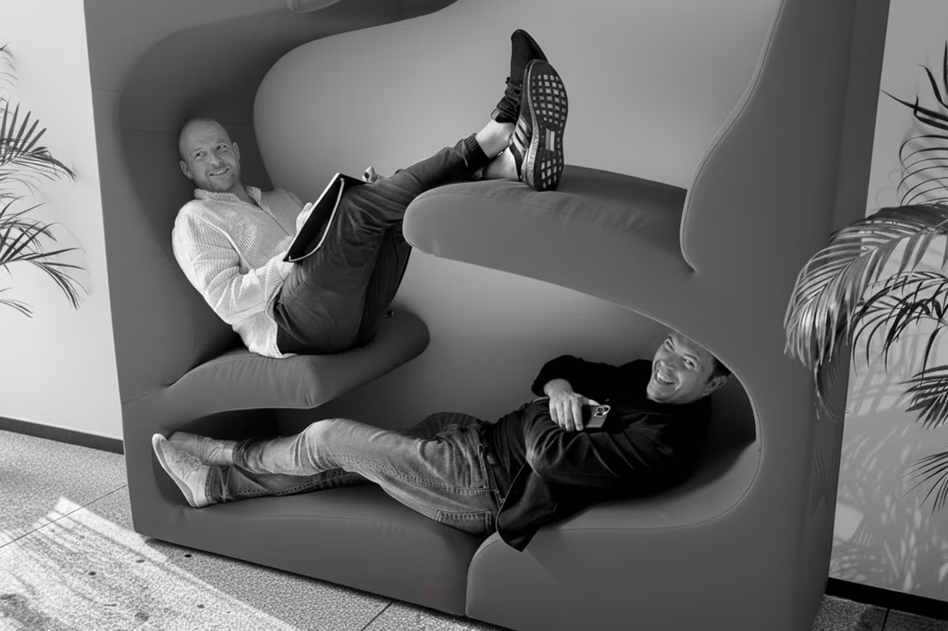 Sculptural lounge pod with two of the four founders reclining in stacked compartments, surrounded by plants and soft natural light