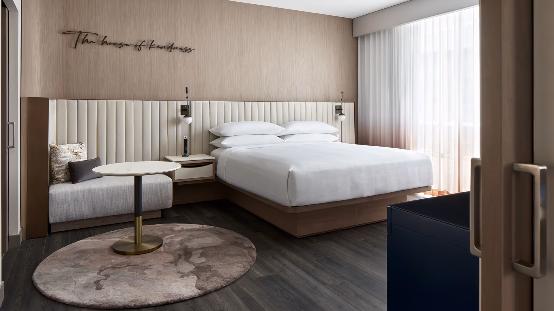 Marriott suite with white linens, built-in nightstands, and a seating nook with gold-base table beneath a wall script reading “The house of kindness”