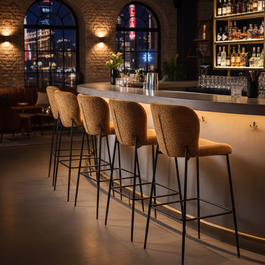 Urban bar interior with 1902 tan Enna upholstered stools, exposed brick walls, and nighttime city view through arched windows
