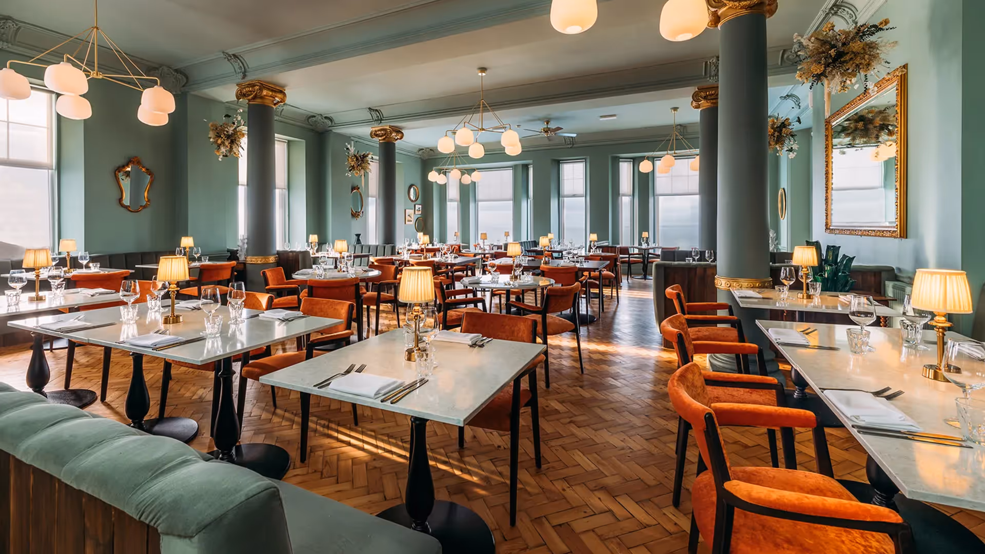 Elegant restaurant interior with marble-topped tables, 1902 Rizo orange upholstered chairs, and teal columns beneath ornate mirrors and chandeliers