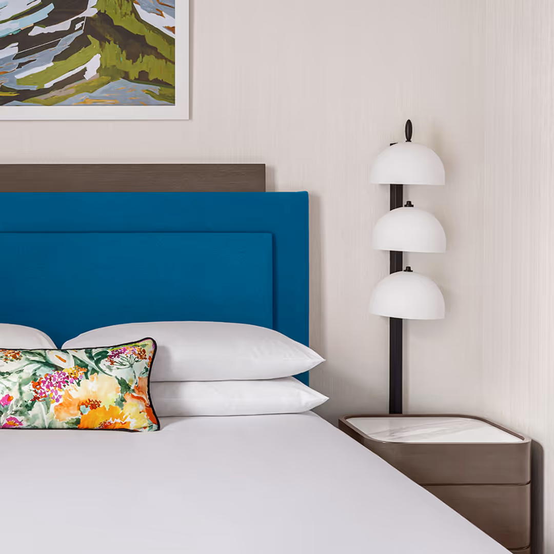 Kimpton hotel suite with teal 1902 case goods, upholstered headboard, floral accent pillow, and three-shade floor lamp beside nightstand and mountain artwork