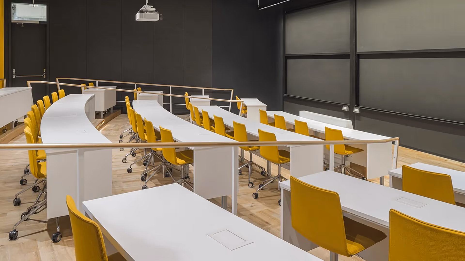Harvard lecture hall with curved white desks, yellow upholstered chairs, and built-in power outlets beneath ceiling-mounted projector