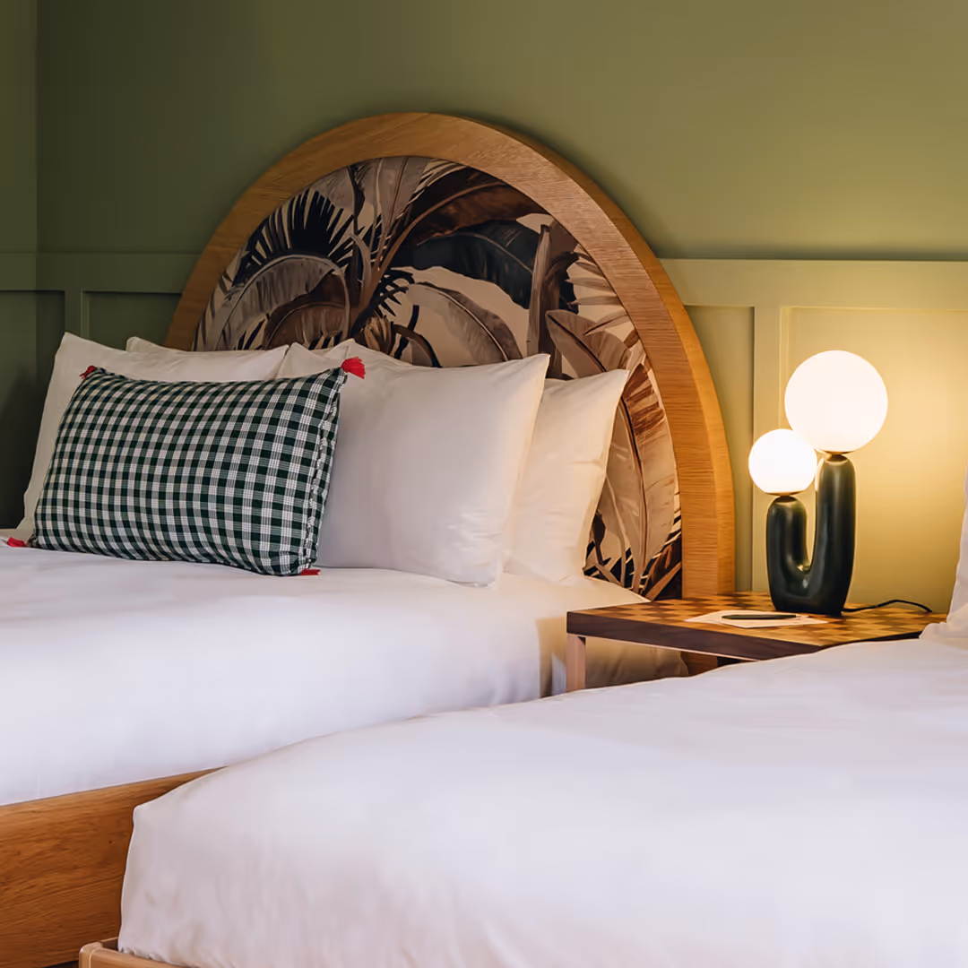 Wayfinder twin bedroom with 1902 case goods, tropical leaf headboard, green accent pillow with tassels, and soft green panelled wall with spherical lamp