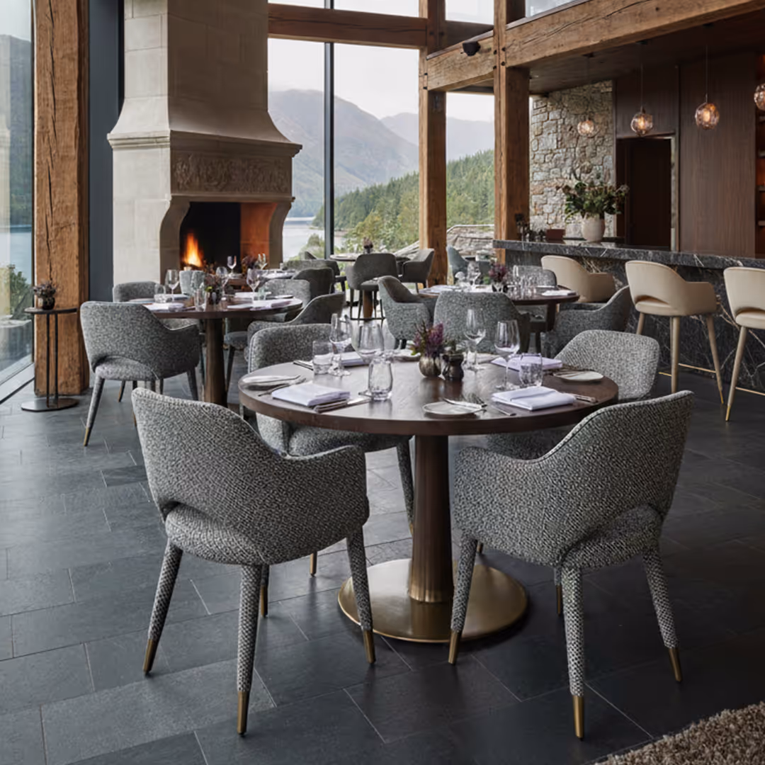 Scenic restaurant interior with lake and mountain views, stone fireplace, and round tables set beneath pendant lights and 1902 Savoca and Sesto brass-tipped seating