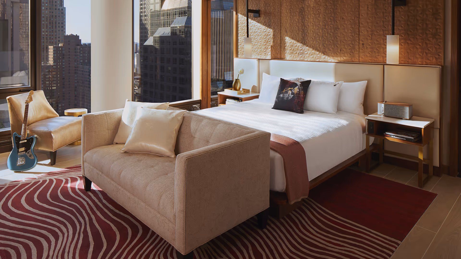 Hard Rock NYC guestroom with 1902 case goods, white linens, beige sofa, and red patterned rug featuring a guitar on a chair and city view through large windows
