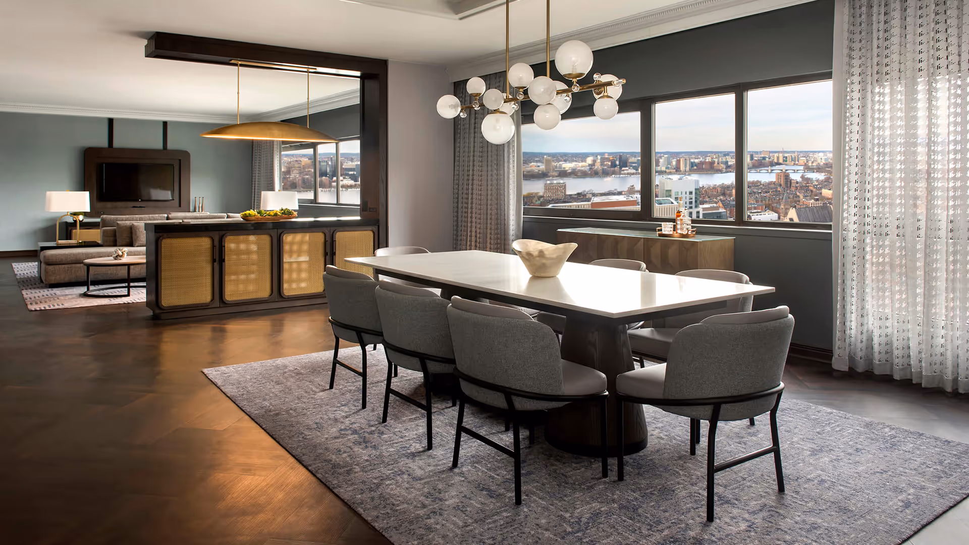 Marriott suite with 1902 case goods and seating, glossy table, light grey chairs, and spherical chandelier beside city-view windows and gold pendant light