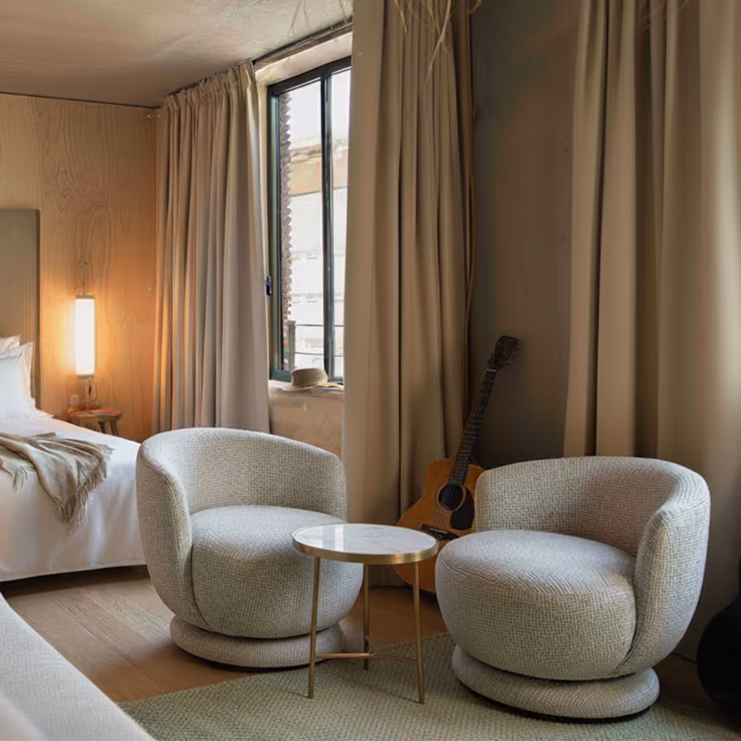 Cozy hotel guestroom with two 1902 Jett rounded armchairs, marble side table, acoustic guitar, and warm wood panelling