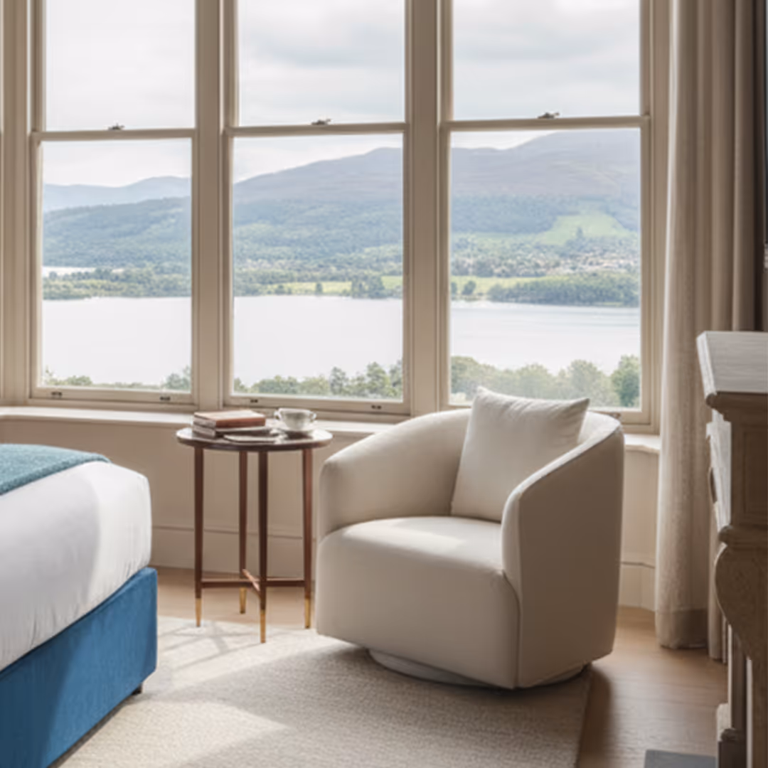 Luxury hotel suite with 1902 Zaria swivel accent chair, wood side table, lake view through panoramic window, and blue upholstered bed