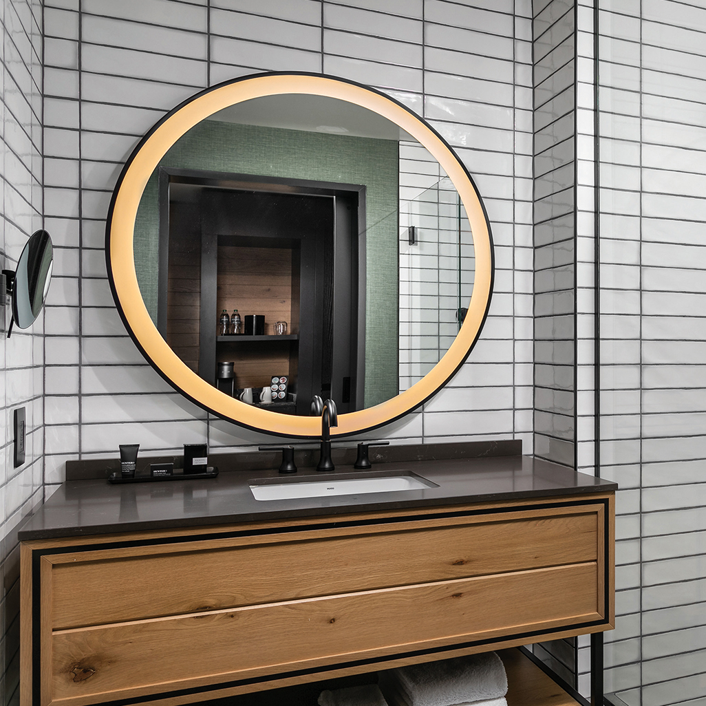 Stylish bathroom at Curio by Hilton featuring a glowing round mirror, sleek wood vanity, and crisp tiled walls.