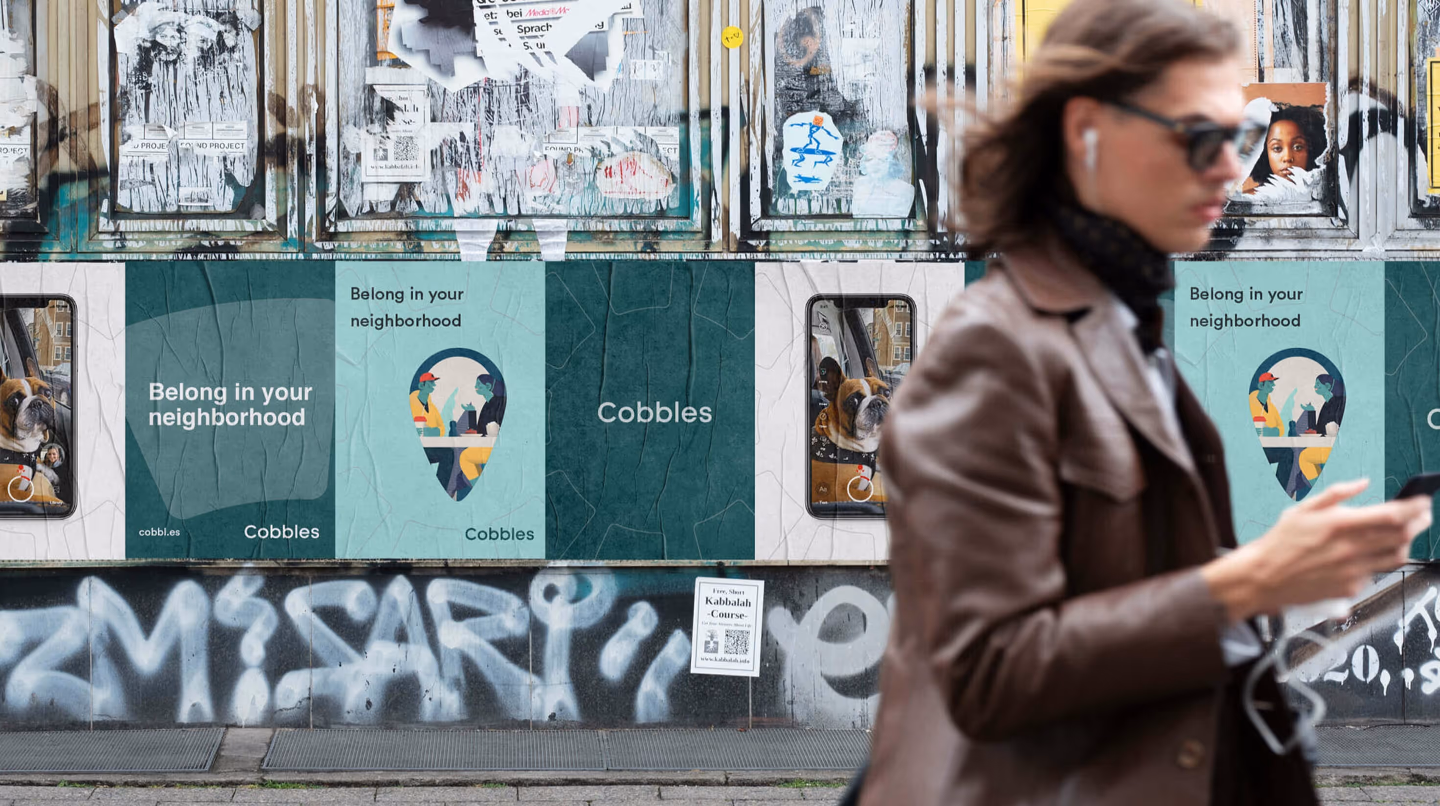 Street posters for Cobbles with the slogan ‘Belong in your neighborhood,’ next to a person walking past using a phone.