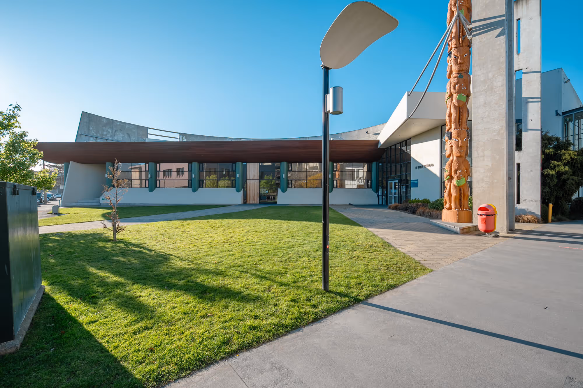 Outside view of Ara Te Puna, with grass and a Māori equivalent to a totem pole which is called is a pouwhenua (or pou)