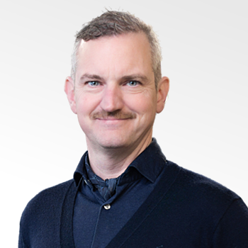 Smiling middle-aged man with short gray hair and a mustache wearing a dark sweater over a collared shirt.