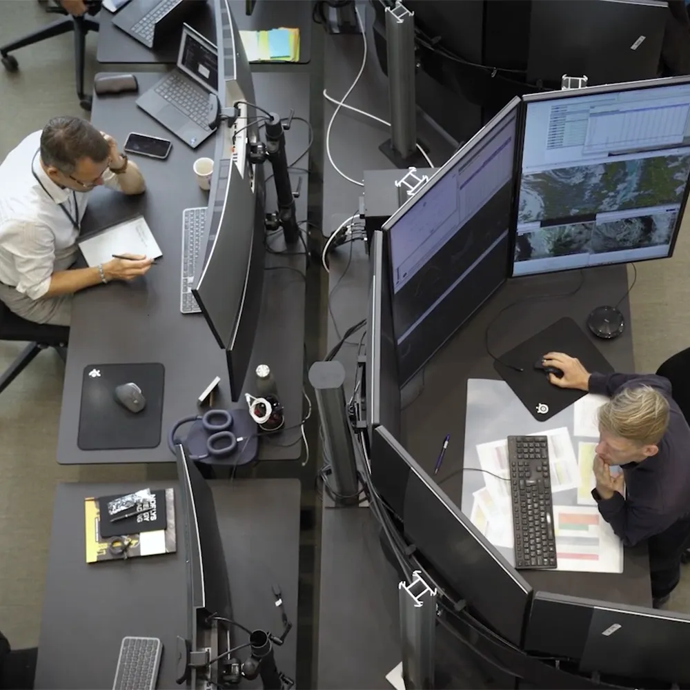 Two men working at separate desks with multiple monitors, laptops, and office supplies in a modern workspace.