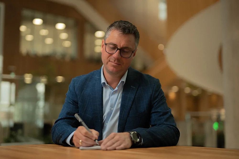 Man wearing glasses and a blue blazer writing with a pen at a wooden table in an office setting.