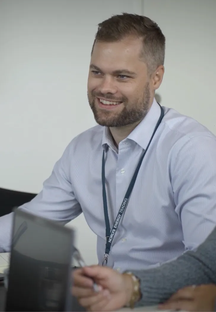 Smiling man in a light blue shirt wearing a lanyard that reads 'NORLYS ENERGY TRADING' seated at a table in an office setting.