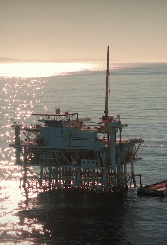 Offshore oil platform in calm sea with sun reflecting on the water and a boat nearby.