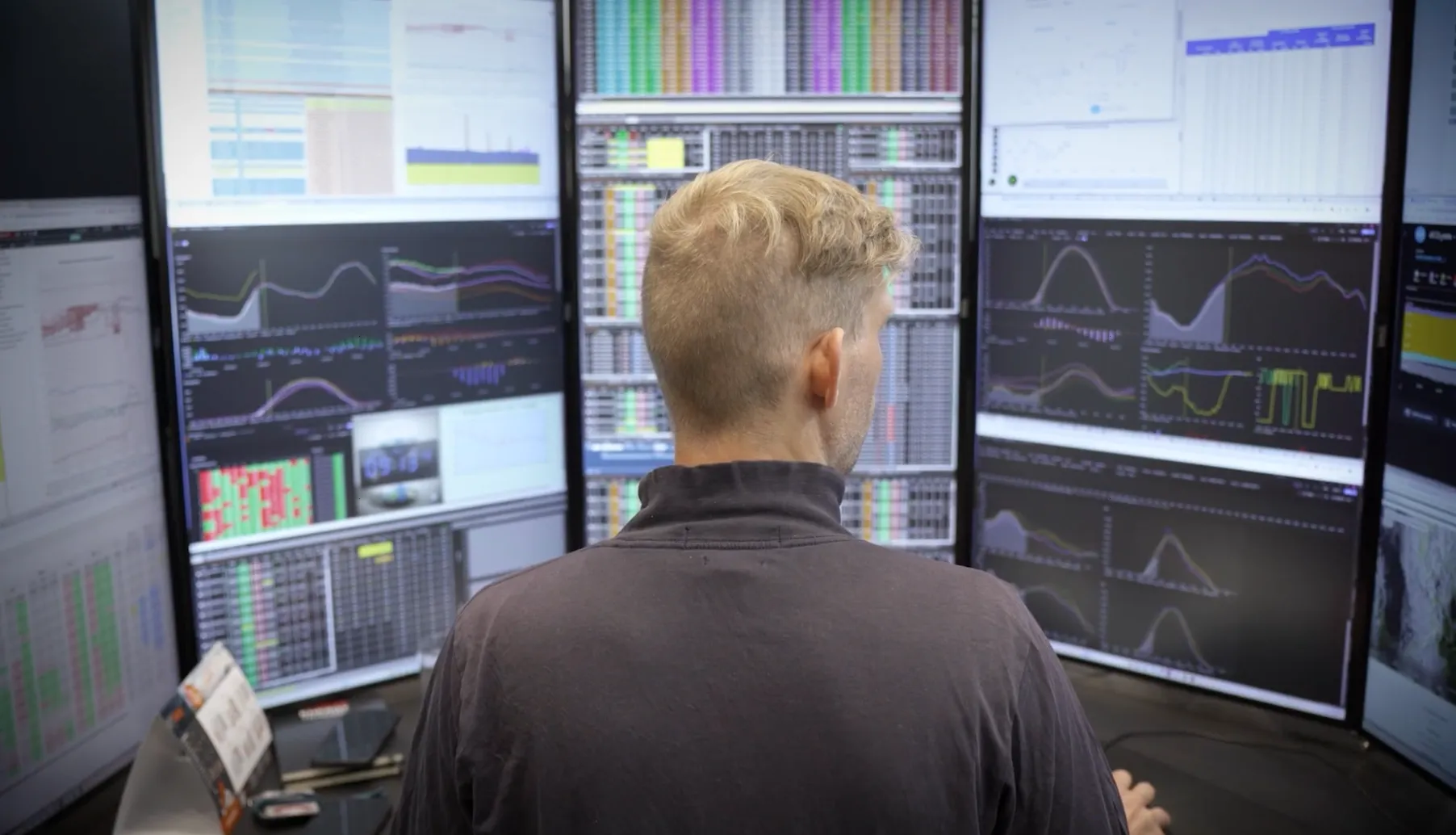 Person with short blonde hair sitting in front of multiple large monitors displaying financial charts and data.