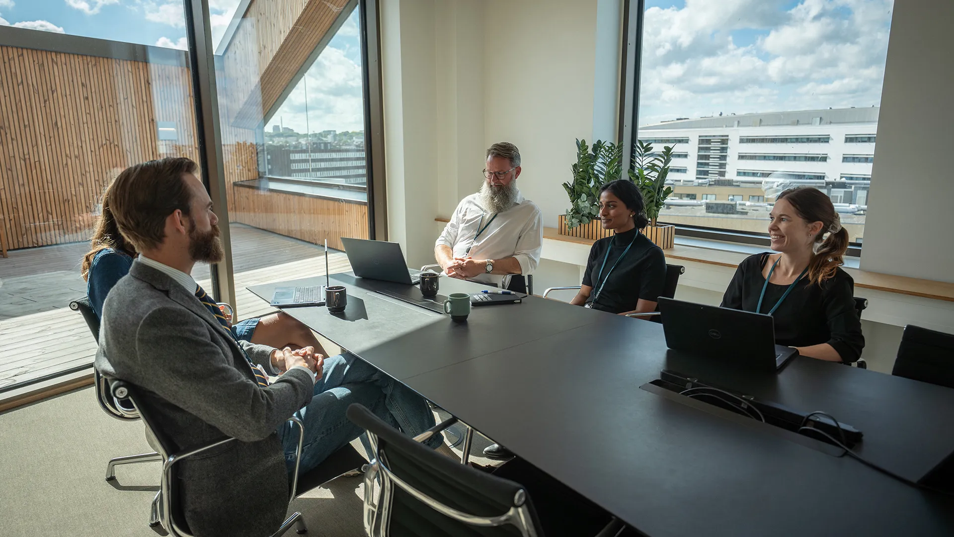 Five professionals seated around a conference table in a bright office with large windows, laptops, and coffee mugs during a meeting.