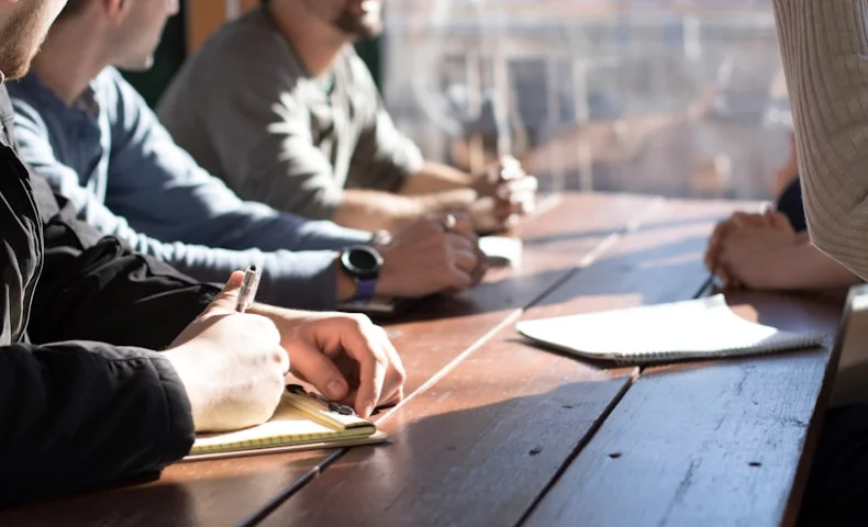 People sitting at a wooden table in a meeting, writing notes in notebooks during discussion.