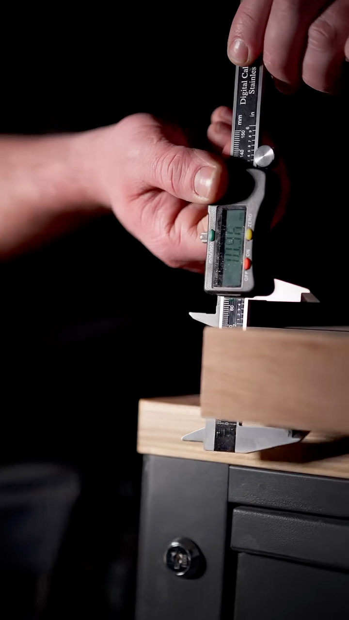 Main tenant un pied à coulisse numérique mesurant l'épaisseur d'une planche en bois clair sur un établi noir.
