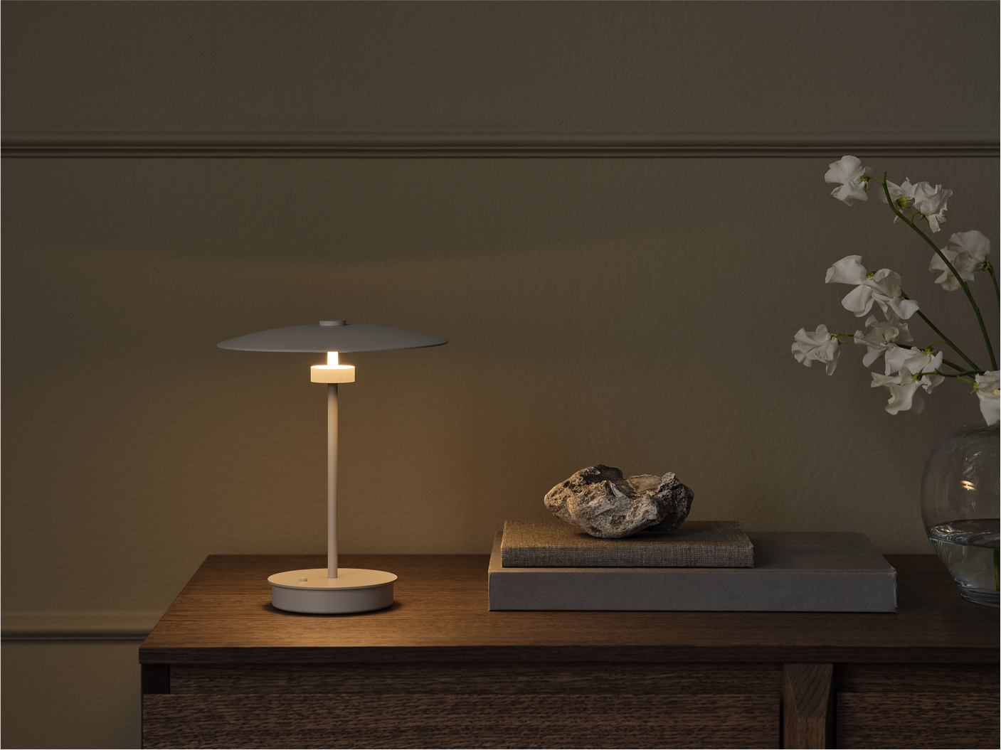 Lampe de table blanche allumée sur une commode en bois avec deux livres et une pierre décorative à côté de fleurs blanches dans un vase en verre.