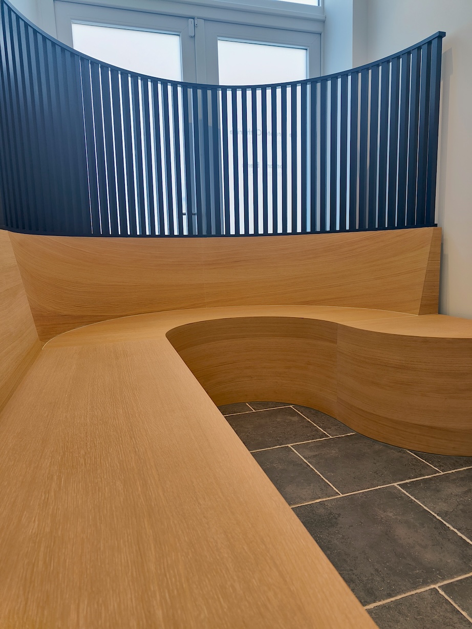 Banquette en bois courbée autour d'un sol carrelé gris avec un garde-corps en lattes noires.
