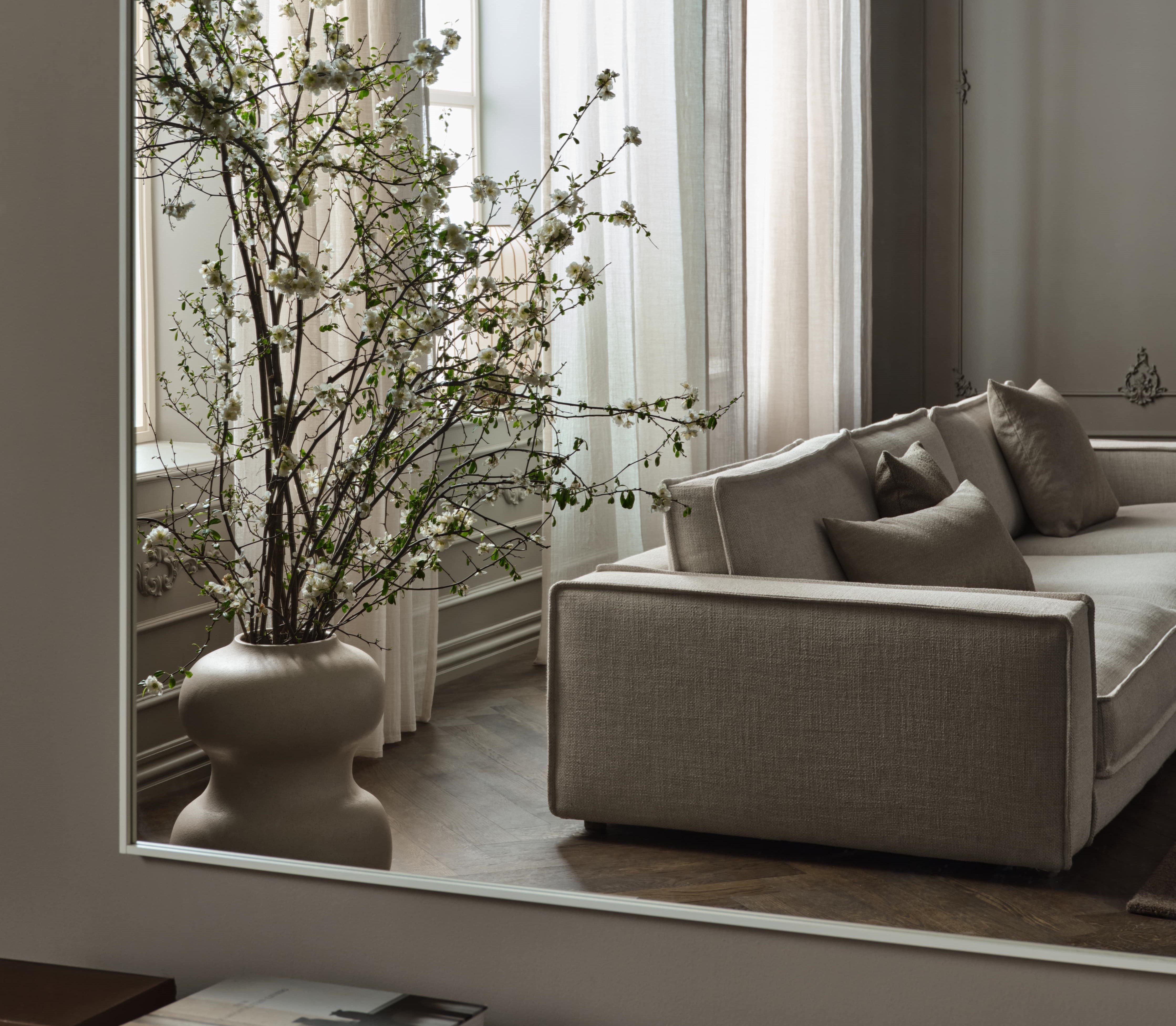 Intérieur élégant avec canapé gris clair et grand vase beige contenant des branches fleuries blanches devant une fenêtre avec rideaux translucides.