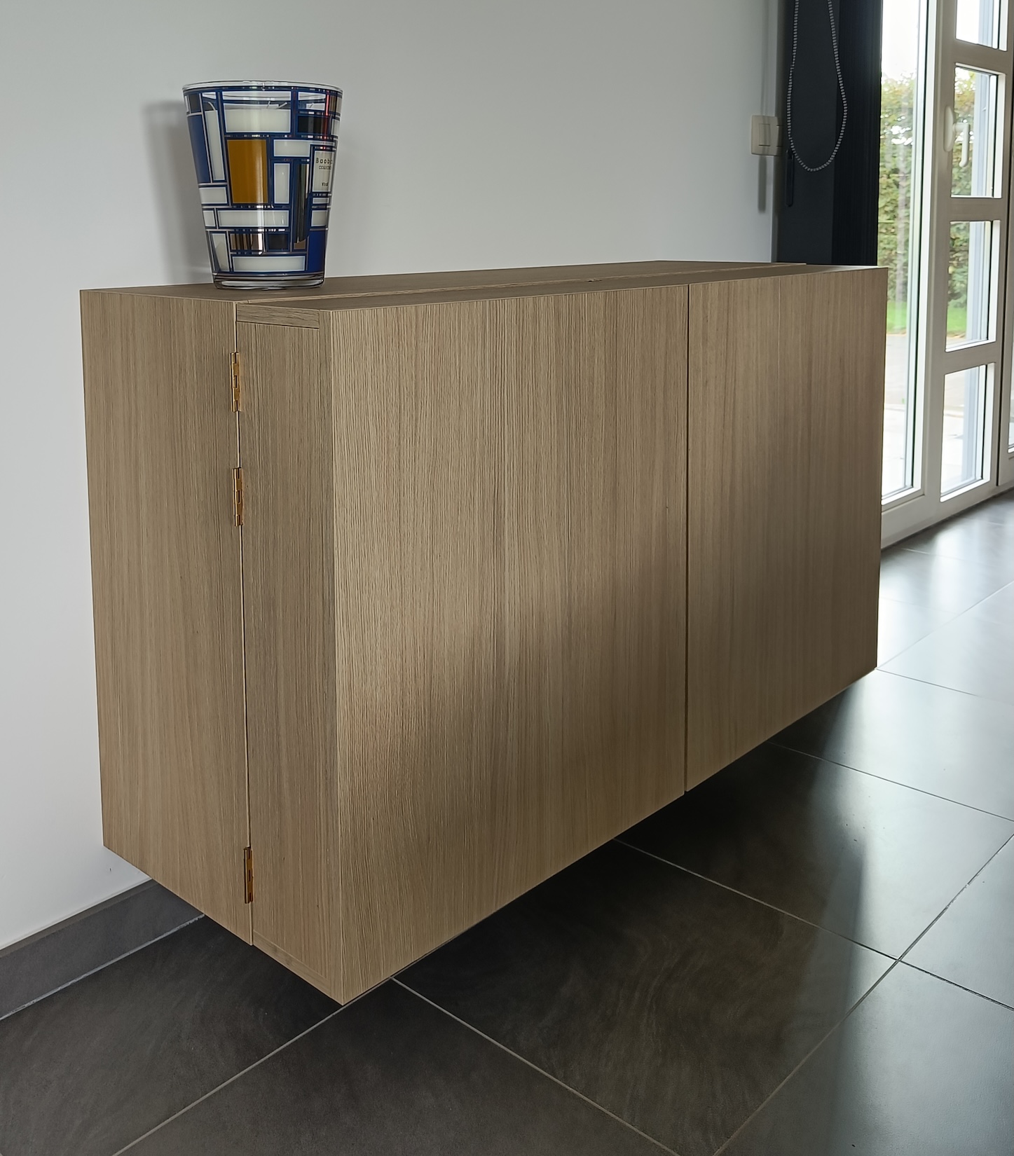 Meuble suspendu en bois clair avec un vase décoratif bleu, blanc et jaune posé dessus, placé dans une pièce lumineuse avec un sol carrelé noir.