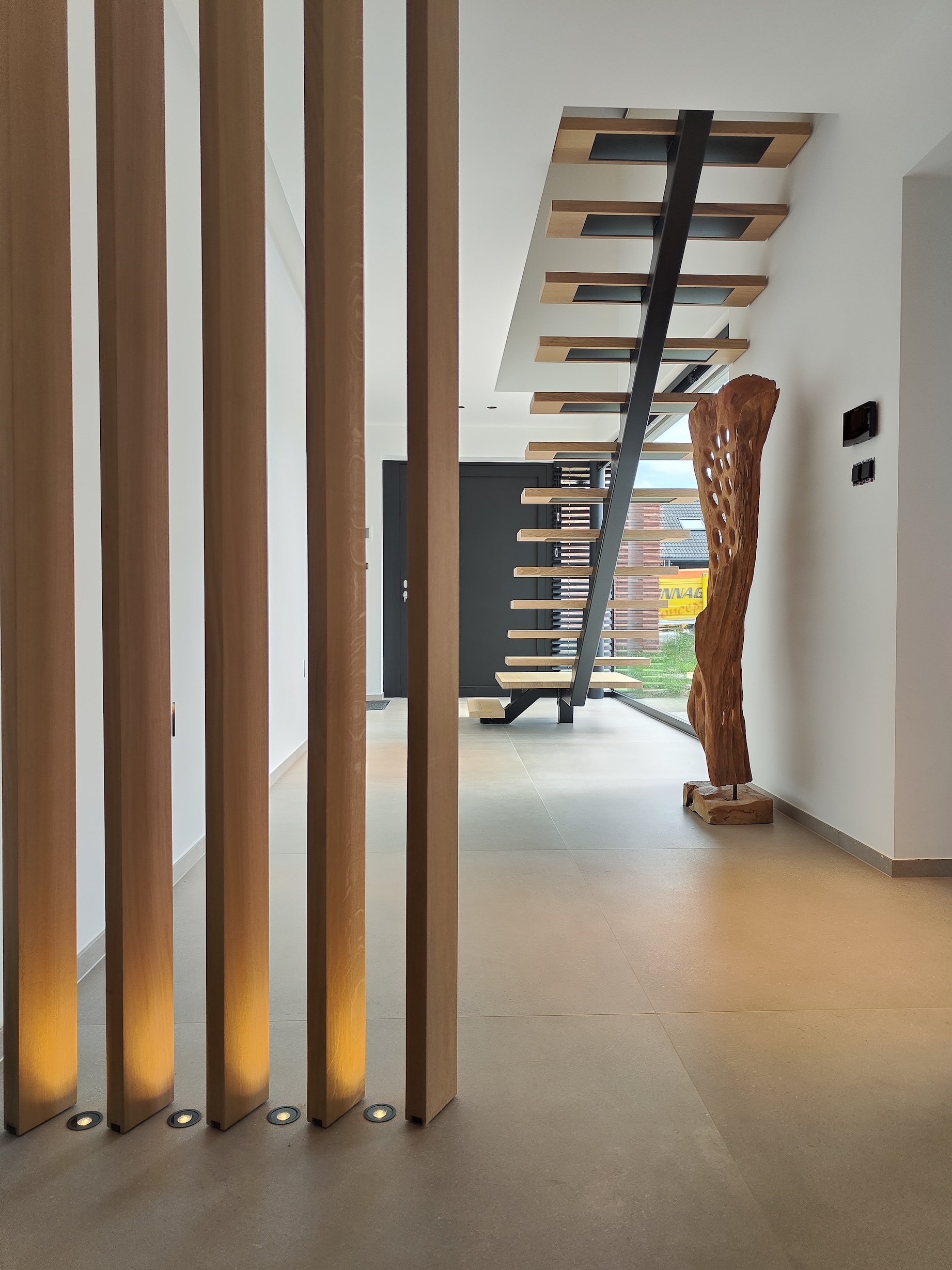 Intérieur moderne avec des piliers en bois éclairés au sol, un escalier flottant en bois et métal, et une sculpture en bois près d'une grande fenêtre.