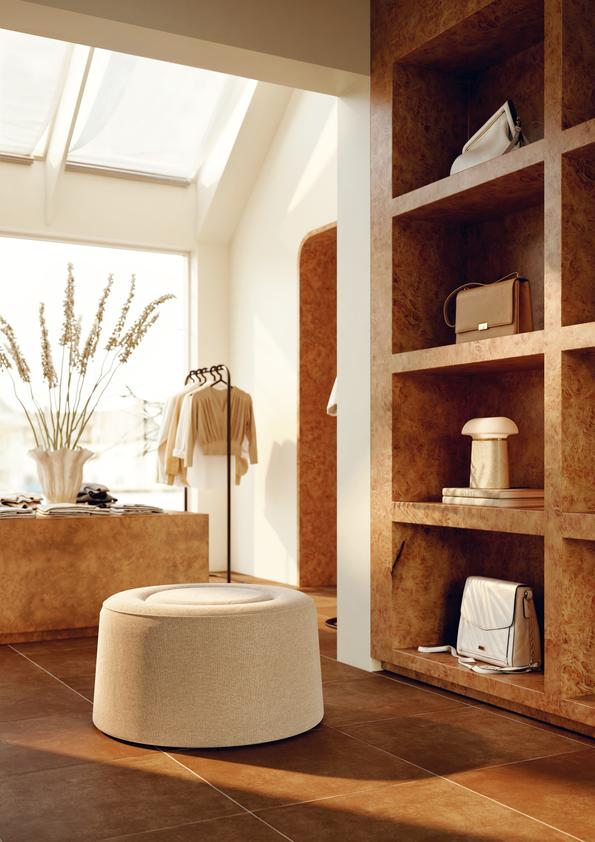 Intérieur élégant d'une boutique avec un pouf rond beige au centre, des étagères en bois avec sacs à main, des vêtements suspendus, et une grande fenêtre laissant entrer la lumière naturelle.