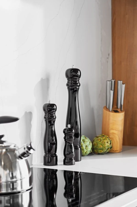 Deux moulins à poivre noirs de tailles différentes, un plus grand au centre et un plus petit à droite, posés sur un plan de travail blanc avec un set de couteaux en bois et deux artichauts en arrière-plan.
