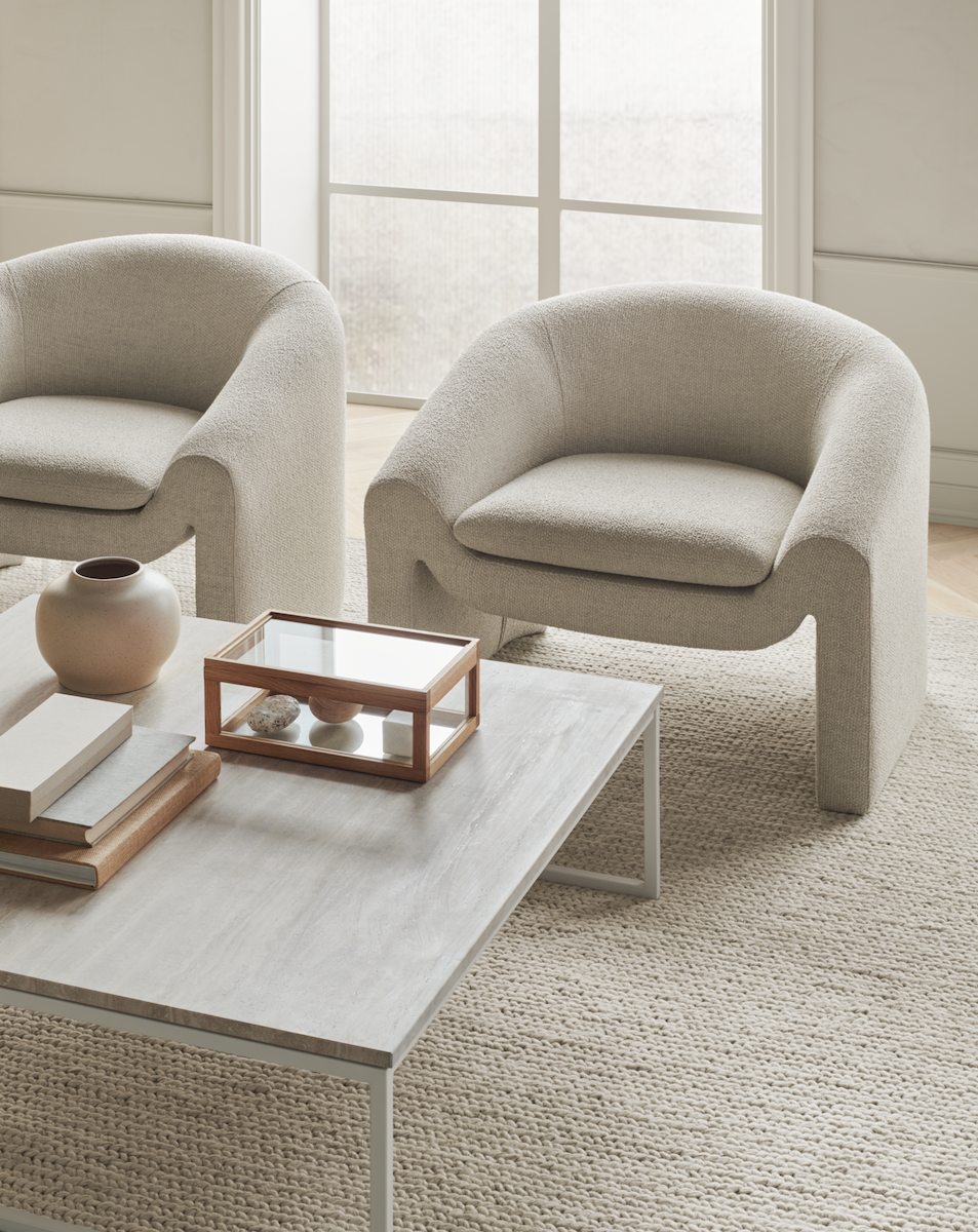 Deux fauteuils beige en tissu avec une table basse en pierre claire décorée d'un vase, de livres et d'un coffret en bois et verre dans un salon lumineux à tapis tissé.